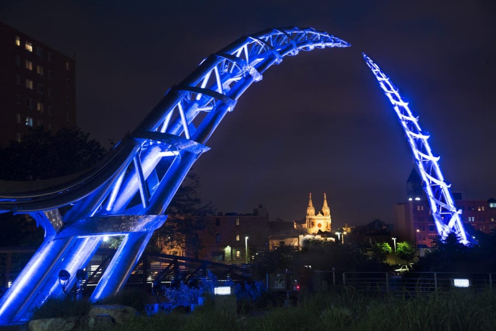 Metal twisting arc sculpture over a city lit up blue at night